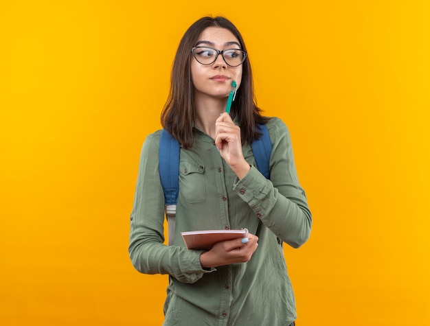 Beeindruckt aussehende junge Schulfrau mit Rucksack mit Brille, die ein Notizbuch mit Stift hält