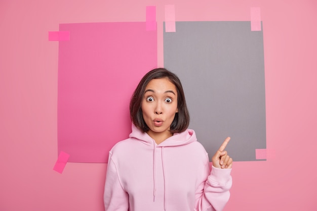 Beeindruckte emotionale junge Asiatin keucht von Schockpunkten mit dem Finger nach oben zeigt etwas unerwartetes Banner nach oben gekleidet in Hoodie-Posen gegen rosa Wand schlägt Sonderverkauf vor