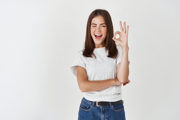 Beeindruckte moderne Frau im T-Shirt, nicken akzeptierend, zwinkernde Show okay, Genehmigungsschild, weiße Wand stehen.