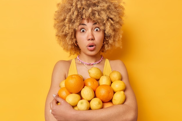 Foto beeindruckte schockierte frau mit lockigen blonden haaren umarmt haufen saftiger frischer orangen und zitronen hält den mund vor wundern geöffnet, die über leuchtend gelbem hintergrund isoliert sind zitrusfrüchte voller vitamine