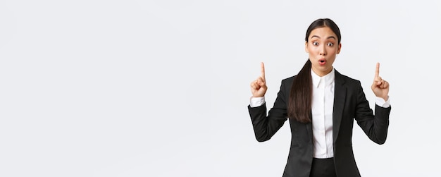 Beeindruckte und aufgeregte asiatische Geschäftsfrau im Anzug diskutieren große Neuigkeiten, die eine Ankündigung zeigen und erstaunt sagen, wow, während sie mit dem Finger nach oben auf weißen Hintergrund zeigen