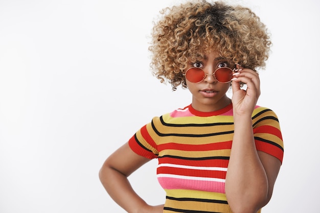 Beeindruckte und erstaunte attraktive, stylische Afroamerikanerin mit blonden Locken in stylischem 90er Jahre gestreiftem Top und cooler Sonnenbrille, die fragend und unsicher in die Kamera schaut, die über weißer Wand posiert