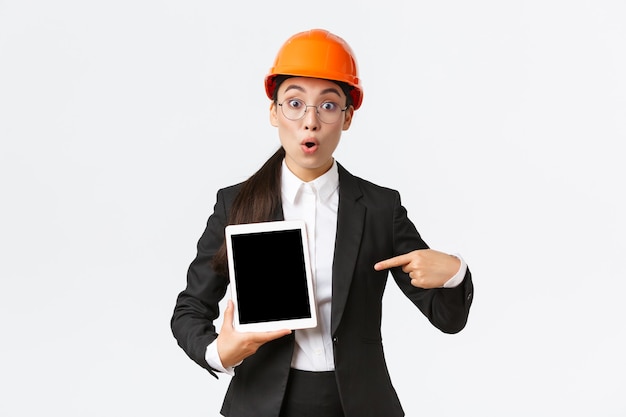 Beeindruckte und überraschte asiatische Ingenieurin, Technikerin im Industriegebiet tragen Schutzhelm und Business-Anzug, zeigen mit dem Finger auf die Gewinntabelle, Diagramm auf dem digitalen Tablet-Bildschirm, sehen erstaunt aus