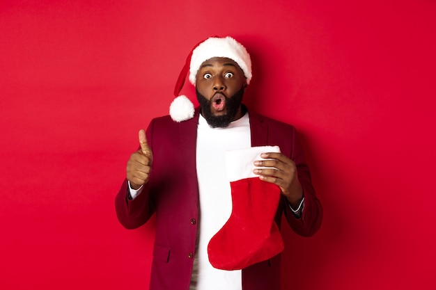 Beeindruckter afroamerikanischer Mann, der Weihnachtssocke mit Geschenken hält, Daumen nach oben zeigt, überrascht aussieht und über rotem Hintergrund steht.