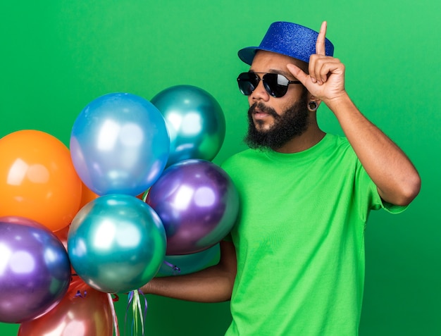 Beeindruckter junger Afroamerikaner mit Partyhut und Brille mit Ballons zeigt nach oben