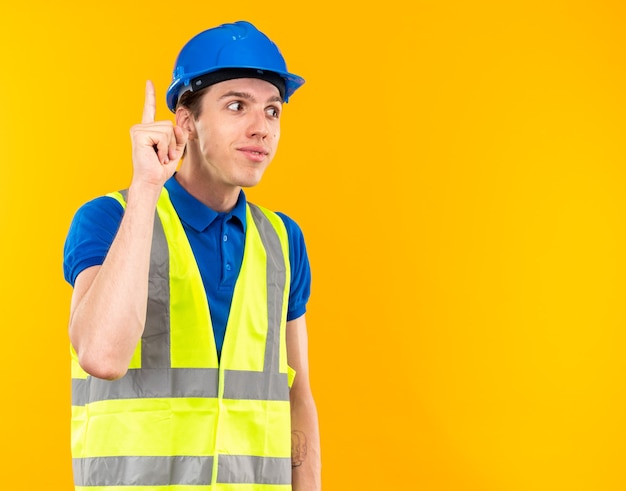 Beeindruckter junger Baumeister in Uniform zeigt nach oben isoliert auf gelber Wand mit Kopierraum
