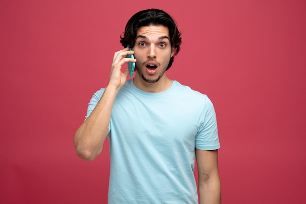 Beeindruckter junger gutaussehender mann, der in die kamera schaut, während er am telefon spricht, isoliert auf rotem hintergrund