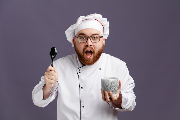 Beeindruckter junger Koch mit einheitlicher Brille und Mütze, der Löffel und Schüssel zeigt, die isoliert auf violettem Hintergrund in die Kamera blickt