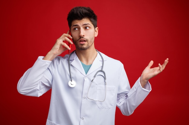 Beeindruckter junger männlicher Arzt, der eine medizinische Uniform und ein Stethoskop um den Hals trägt und eine leere Hand zeigt, die am Telefon spricht und die Seite isoliert auf rotem Hintergrund betrachtet