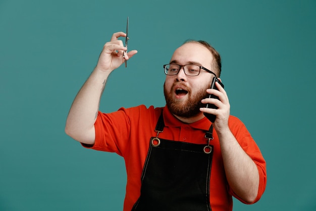 Beeindruckter junger männlicher Friseur mit Brille, rotem Hemd und Friseurschürze, der auf die Seite blickt, die eine Schere hochhebt und am Telefon isoliert auf blauem Hintergrund spricht