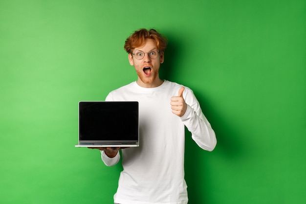 Beeindruckter kaukasischer Mann mit roten Haaren und Bart, der leeren Laptop-Bildschirm und Daumen hoch zeigt, erstaunt in die Kamera schaut und etwas online lobt, grüner Hintergrund.
