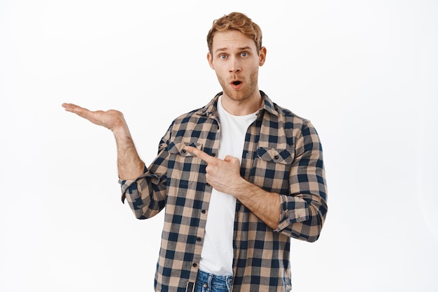 Beeindruckter Kerl, der auf eine offene Hand zeigt, die einen Gegenstand hält, erstaunt über das Produkt auf seiner Handfläche, das leeren Platz für Ihr Logo oder Objekt zeigt, das auf weißem Hintergrund steht