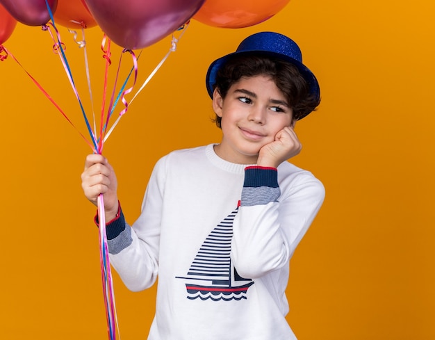 Beeindruckter kleiner Junge mit blauem Partyhut, der Luftballons hält, die Hand auf die Wange legen, isoliert auf oranger Wand