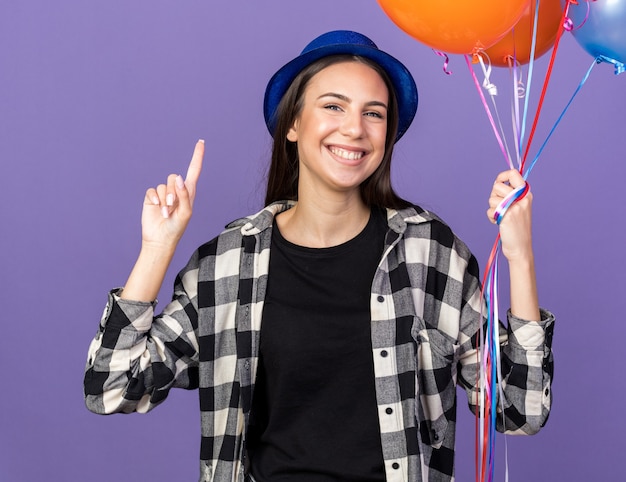 Beeindrucktes junges schönes Mädchen mit Partyhut, das Luftballons hält, zeigt nach oben isoliert auf blauer Wand