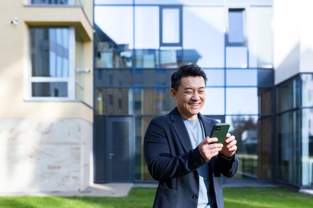 Beendeter asiatischer Geschäftsmann feiert Leistung mit Blick auf Smartphone im Freien Erfolgreicher Geschäftsmann feiert Sieg mit Handy draußen Happy Good News Erfolgreicher Deal Startup Bet