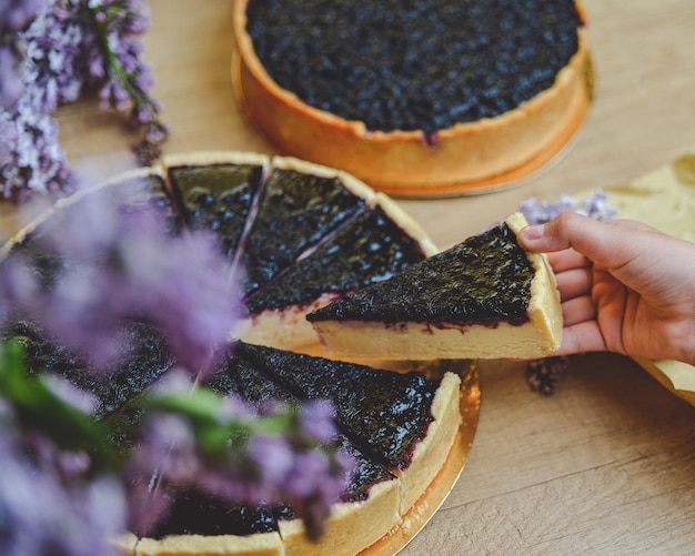 Beeren-Käsekuchen auf einem Holztisch mit lila Blüten um die Draufsicht, Kind hält ein Stück Kuchen