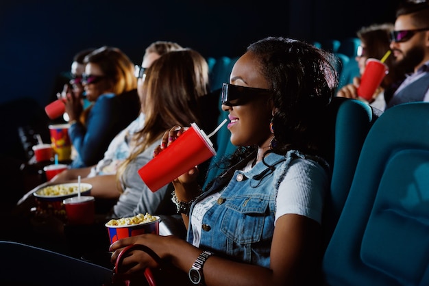 Begeistert, den neuen Film zu sehen. Junge Afrikanerin nippt an ihrem Getränk, während sie einen Film im Kino mit 3D-Brille sieht watching