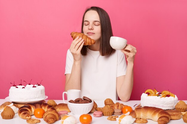 Begeisterte braunhaarige Frau in weißem T-Shirt, die isoliert über rosa Hintergrund am Tisch sitzt und süßes Croissant riecht und eine Tasse Tee hält, hält die Augen geschlossen