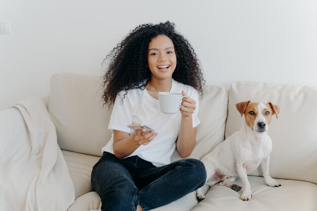 Begeisterte dunkelhäutige Frau posiert in einer modernen Wohnung sitzt auf einem bequemen Sofa mit Haustiergetränken Kaffee nutzt das Mobiltelefon für die Online-Kommunikation und ist gut gelaunt