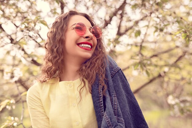 Foto begeisterte frau in stilvollem outfit, die fröhlich lächelt und wegschaut, während sie sich auf unscharfem hintergrund blühender äste im frühlingsgarten entspannt