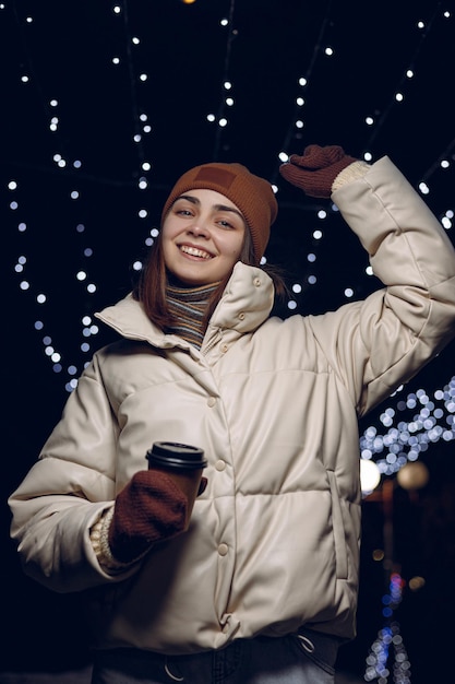 Begeisterte Frau in warmer Oberbekleidung mit einer Tasse Kaffee, die im Winter auf der Stadtstraße tanzt und Spaß hat