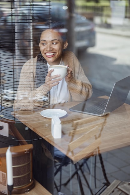 Begeisterte, glückliche Afrikanerin arbeitet am Laptop, lächelt freudig und betrachtet vor dem Fenster Cappuccino