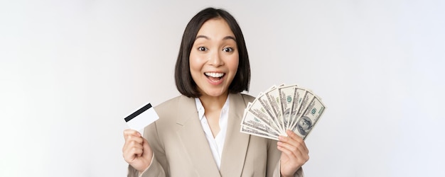 Begeisterte junge asiatische Geschäftsfrau, die Gelddollar und Kreditkarte zeigt, die über weißem Hintergrund stehen