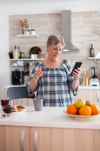Begeisterte Seniorin spricht über die Handy-Webcam mit der Familie, die während des Frühstücks in der Küche sitzt und lächelt. Authentische ältere Person mit moderner Smartphone-Internettechnologie.