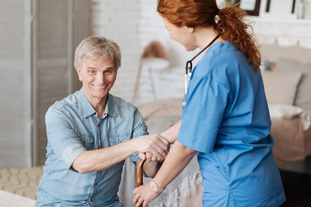 Begeisterte Stimmung. Fröhlicher, lebhafter älterer Herr, der jede Woche von einer Krankenschwester besucht wird, um sich während seiner Behandlung untersuchen zu lassen und Hilfe zu leisten