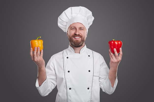 Begeisterter bärtiger männlicher Koch in weißer Kochuniform, der mit gelben und roten frischen Paprika auf grauem Hintergrund im Studio steht und in die Kamera schaut