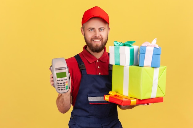 Begeisterter Kuriermann, der einen Stapel Geschenkboxen und ein POS-Terminal für Online-Zahlungen hält