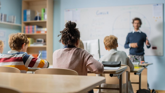 Begeisterter Lehrer erklärt Lektionen und stellt Fragen aus einem Klassenzimmer voller brillanter, vielfältiger Kinder in der Grundschule Eine Gruppe intelligenter, multiethnischer Kinder lernt Naturwissenschaften und kreatives Denken