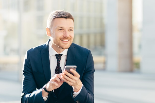 Begeisterter, nachdenklicher, eleganter männlicher Marketing-Händler im formellen schwarzen Anzug verwendet ein modernes Handy für die Suche auf der Website in Internet-Typen Textnachrichten blickt nachdenklich in die Ferne Geschäftskonzept