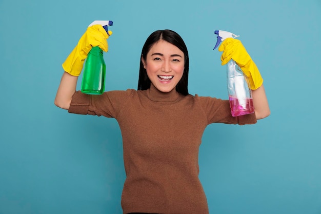 Begeistertes, frohes asiatisches Hausmädchen, das die Arme hebt und zwei Waschmittelsprays in den gelben Handschuhen hält, Hausfrauenarbeiterin, Reinigungshauskonzept, lächelnd fröhlich und positiv von der täglichen Arbeit