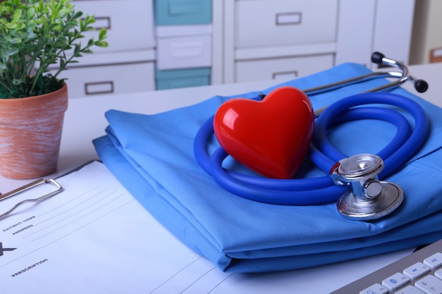 Behandeln Sie Mantel mit medizinischem Stethoskop und rotem Herzen auf dem Schreibtisch