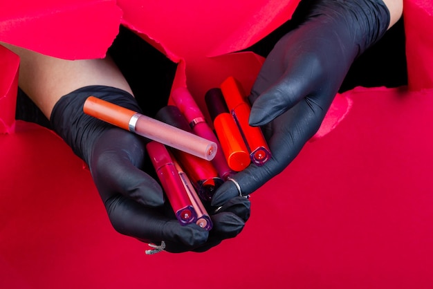 Behandschuhte Hände, die einen Haufen verschiedener Lipgloss-Sticks halten, die eine rote Papierwand durchbrechen