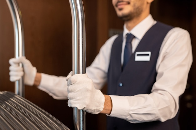 Foto behandschuhte hände eines jungen portiers in uniform, der den wagen mit koffern von gästen schiebt, während er sich vor der kamera entlang des korridors im hotel bewegt?