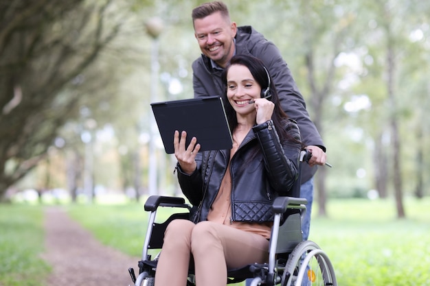 Behinderte Frau im Rollstuhl und Mann, die auf digitalem Tablet im Park sprechen, Fernarbeit für Menschen