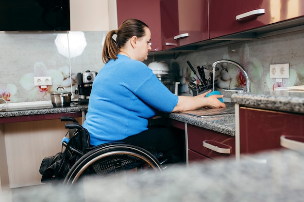 Behinderte junge Frau im Rollstuhl, die Geschirr in der speziell ausgestatteten Küche spült