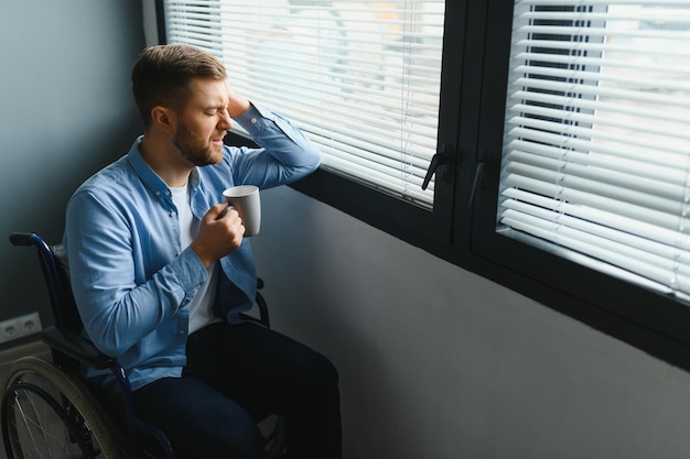 Behinderte Person sitzt im Rollstuhl am Fenster Ernster trauriger kaukasischer Mann in lässiger Kleidung und Blick auf den großen Panoramablick im hellen modernen Wohnzimmer oder Krankenhaus