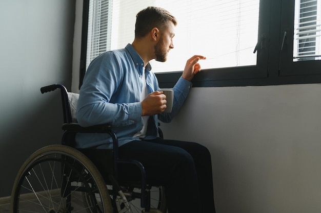 Behinderte Person sitzt im Rollstuhl am Fenster Ernster trauriger kaukasischer Mann in lässiger Kleidung und Blick auf den großen Panoramablick im hellen modernen Wohnzimmer oder Krankenhaus