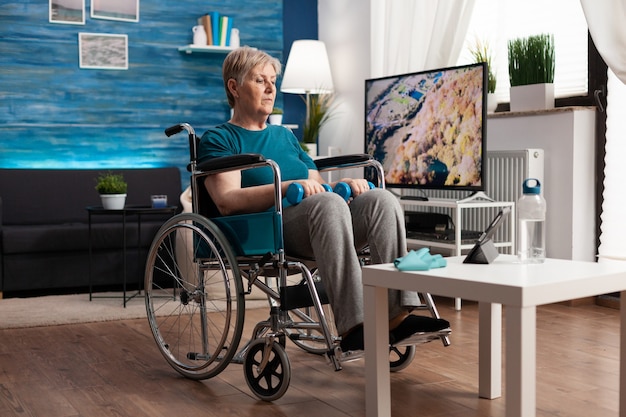Behinderte Seniorin im Rollstuhl, die sich gymnastisches Online-Video auf einem Tablet-Computer ansieht und Körpermuskeln mit Hanteln trainiert