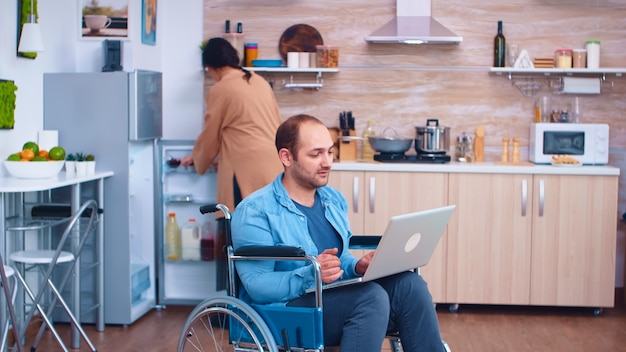 Behinderter Geschäftsmann im Rollstuhl, der am Laptop arbeitet, während Frau in der Küche kocht. Geschäftsmann mit Lähmung Handicap Behinderung behinderte Schwierigkeiten bei der Arbeit nach einem Unfall mit Internet