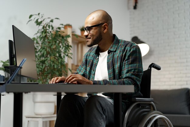 Behinderter junger Afroamerikaner im Rollstuhl mit Computer, während er an seinem Arbeitstisch sitzt