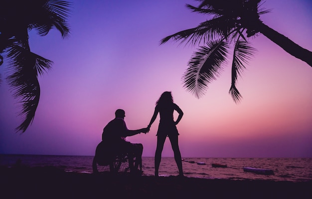 Behinderter Mann im Rollstuhl mit seiner Frau am Strand. Silhouetten bei Sonnenuntergang