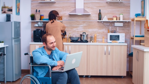 Behinderter Mann winkt auf Videokonferenz mit Laptop in der Küche. Geschäftsmann mit Lähmung Behinderung Behinderung behinderte Schwierigkeiten bei der Arbeit nach einem Unfall mit Internet-Online-Videoanruf conf