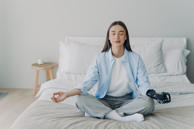 Behindertes Mädchen mit bionischer Armprothese praktiziert Yoga und meditiert in Lotus-Pose auf dem Bett zu Hause