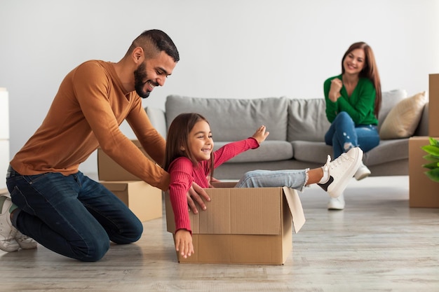 Bei Neue Wohnung. Fröhliche Familie aus dem Nahen Osten, die Spaß hat und den Umzugstag feiert, aufgeregter Vater, der seine kleine Tochter im Karton reitet, glückliche Mutter, die auf der Couch in unscharfem Hintergrund sitzt