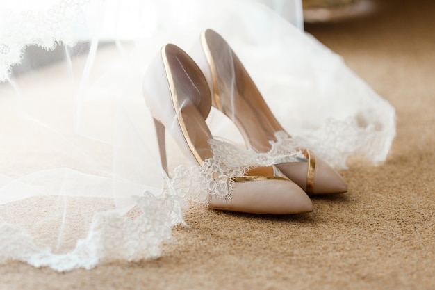 Beige Brautschuhe stehen auf dem Boden, bedeckt mit einem weißen Schleier mit Spitze