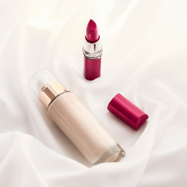 Beigefarbene Cremeflasche Make-up-Flüssigkeitsgrundlage und roter Lippenstift auf Seidenhintergrund-Kosmetikprodukten als luxuriöses Schönheitsmarken-Urlaubsdesign